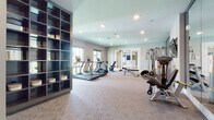 This single-floor fitness center is thoughtfully designed to accommodate a full range of workout routines. The space is divided into distinct functional zones, each outfitted with professional-grade equipment. The main cardio and strength area features treadmills, an elliptical, a stationary bike, and a multi-station cable machine, complemented by a rack of dumbbells in varying weights. Large wall-mounted mirrors throughout amplify the sense of space and allow users to monitor their form during exercise. A wall-mounted flat-screen TV adds an entertainment element to training sessions. An adjoining room serves as a functional training studio, finished with durable rubberized cork flooring ideal for high-intensity workouts. This area is stocked with medicine balls, stability balls, kettlebells, resistance bands, foam rollers, and additional cable training equipment, along with full-length mirrors. A separate, more serene room offers a calming environment suited for stretching, yoga, or low-impact exercise, featuring neutral-toned walls, a large window with exterior views, and a piece of framed artwork. Recessed lighting throughout the facility creates a clean, modern aesthetic, while the neutral color palette and organized storage solutions contribute to an inviting and functional atmosphere.
