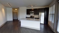 This single-floor residence showcases a well-designed open-concept layout, with the kitchen serving as a central highlight. The kitchen features dark espresso-toned cabinetry that contrasts elegantly with a white subway tile backsplash and light granite countertops. A spacious island with a built-in sink and matching granite surface provides both additional prep space and a natural gathering point. Stainless steel appliances, including a refrigerator, range, and over-the-range microwave, lend a polished, modern aesthetic. Track lighting installed on the ceiling illuminates the kitchen efficiently. Dark hardwood-style flooring extends throughout the open living area, adding warmth and visual continuity to the space. The open sightlines from the kitchen toward the living area create a sense of spaciousness, making the layout practical for everyday living and entertaining. A hallway visible in the background leads toward the entry, featuring a paneled door that hints at a well-organized floor plan. The overall ambiance is contemporary and clean, characterized by a neutral color palette, modern fixtures, and quality finishes throughout.