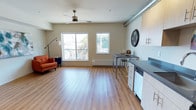 This thoughtfully designed single-floor studio unit blends modern comfort with industrial-inspired character. The open-plan layout seamlessly connects the living and kitchen areas, creating a spacious and versatile environment flooded with natural light from two large windows that frame views of the surrounding neighborhood.

The living area features warm hardwood-style flooring that extends throughout the unit, complemented by neutral beige walls that lend a clean, inviting atmosphere. A ceiling fan with a brushed metal finish adds both function and style overhead, while an exposed cylindrical ventilation duct running along the upper wall reinforces the space's contemporary industrial aesthetic.

The kitchen area is compact yet functional, outfitted with light wood cabinetry, dark countertops, and a built-in dishwasher. A metal-framed bar-height table with stools serves as a convenient dining and workspace. A large decorative wall clock adds a bold, eye-catching accent above the cabinetry.

The living zone is accented by a vibrant orange armchair and a tall arc floor lamp, contributing warmth and personality to the open space. A baseboard heating unit beneath the windows ensures year-round comfort. The combination of clean lines, natural light, and industrial details makes this unit a distinctive and practical living space.