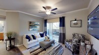 This single-floor residence offers a cohesive, thoughtfully designed layout connecting its key living spaces. The living room serves as the heart of the home, featuring crown molding, a ceiling fan with light fixture, and soft carpet underfoot. A sliding glass door opens to a private balcony, filling the room with natural light. A wall-mounted television and built-in media console anchor the entertainment area on the opposite wall.

The galley-style kitchen is equipped with warm wood cabinetry, dark countertops, and black appliances including a range, microwave, and refrigerator. Dark hardwood-style flooring runs through the kitchen and flows into an adjacent dining area, which comfortably accommodates a round table and chairs.

The bedroom is a calm retreat with plush carpet, a ceiling fan, and a generously sized window that brings in natural light and outdoor views. The full bathroom includes a tub-shower combination, a vanity with warm wood cabinetry, a large mirror with overhead lighting, dark hardwood-style flooring, and direct access to a spacious walk-in closet. The overall palette of soft grays and warm wood tones creates a consistent, welcoming ambiance throughout.
