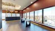 Located at 345 St Peter St in downtown Saint Paul, this striking amenity lounge occupies a single floor of a high-rise building, offering an immersive blend of bold interior design and breathtaking panoramic views of the Saint Paul skyline. Floor-to-ceiling windows wrap the main gathering area, flooding the space with abundant natural light while framing iconic landmarks including the Minnesota State Capitol dome. Large-format porcelain tile flooring runs throughout, providing a sleek, cohesive foundation. The centerpiece of the lounge is a dramatic statement chandelier composed of cascading multicolored metallic fragments, suspended above the main seating area and commanding immediate visual attention. A sculptural navy blue sofa anchors a plush lounge zone set atop a vibrant floral area rug, complemented by a bold dining table with architectural curved legs. A built-in wet bar with navy fluted cabinetry, gold accents, and open shelving serves as both a functional and decorative focal point. A wood-paneled corridor leads to a cozy private nook featuring a curved, upholstered booth with vertical channel tufting and globe pendant lighting framed in gold. Deep terracotta-toned accent walls, gold trim details, and curated lighting throughout create a warm, sophisticated, and distinctly contemporary ambiance ideal for social gatherings and events.