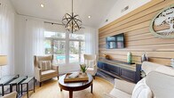 This single-floor amenity space blends coastal-inspired design with modern functionality, creating a welcoming environment that is both practical and visually impressive. The entry area sets an immediate tone with soaring ceilings, crisp white walls, sheer floor-length curtains, and light-toned hardwood-style flooring that extends throughout. Oversized glass double doors flood the space with natural light and provide a seamless connection to the lush outdoor surroundings.

Adjacent to the entry, a business center offers a long built-in workstation with multiple computer stations, a marble-topped communal table with upholstered bar seating, and a statement rectangular chandelier overhead. Tall clerestory windows bathe the area in abundant daylight, while the soaring ceiling height adds a sense of openness and grandeur.

The lounge area is a standout feature, characterized by a dramatic horizontal wood-planked accent wall paired with a built-in navy media console and wall-mounted flat-screen television. A sculptural orb chandelier anchors the seating arrangement, which includes cream wingback chairs, a woven jute rug, and a round dark-wood coffee table. Large picture windows frame direct views of an outdoor pool and tropical landscaping, enhancing the bright, resort-like ambiance throughout this thoughtfully designed space.