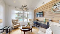 This single-floor amenity space blends coastal-inspired design with modern functionality, creating a welcoming environment that is both practical and visually impressive. The entry area sets an immediate tone with soaring ceilings, crisp white walls, sheer floor-length curtains, and light-toned hardwood-style flooring that extends throughout. Oversized glass double doors flood the space with natural light and provide a seamless connection to the lush outdoor surroundings.

Adjacent to the entry, a business center offers a long built-in workstation with multiple computer stations, a marble-topped communal table with upholstered bar seating, and a statement rectangular chandelier overhead. Tall clerestory windows bathe the area in abundant daylight, while the soaring ceiling height adds a sense of openness and grandeur.

The lounge area is a standout feature, characterized by a dramatic horizontal wood-planked accent wall paired with a built-in navy media console and wall-mounted flat-screen television. A sculptural orb chandelier anchors the seating arrangement, which includes cream wingback chairs, a woven jute rug, and a round dark-wood coffee table. Large picture windows frame direct views of an outdoor pool and tropical landscaping, enhancing the bright, resort-like ambiance throughout this thoughtfully designed space.