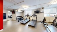 Located at 12-24 Leith Hill Road in Toronto, Ontario, this exercise room offers a well-equipped fitness facility designed to support a variety of workout routines. The space is organized across a single floor and makes efficient use of its footprint, providing a comfortable and functional environment for residents or users.

The room features a clean, neutral aesthetic with white walls accented by a bold red structural column near the entrance, which adds a striking visual contrast. The flooring throughout is a durable, speckled vinyl composition tile, well-suited for high-traffic fitness use. Circular flush-mount light fixtures provide bright, even overhead illumination, while windows along one wall allow natural light to filter in.

Cardio equipment is a highlight of the space, with multiple treadmills positioned toward the center of the room alongside a rowing machine and a stationary bike. A wall-mounted flat-screen television serves the cardio area. A dedicated strength and flexibility zone includes a dumbbell rack with a full range of free weights, a weight bench, and a set of stability balls on a storage rack. A large mirror along one wall enhances the sense of space and supports proper form during exercise.

Additional amenities include a wall-mounted water fountain with bottle-filling capability, a fire extinguisher, a mini-split air conditioning unit for climate control, and posted safety information. The space is thoughtfully arranged to maximize usability while maintaining clear sightlines throughout.