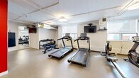 Located at 12-24 Leith Hill Road in Toronto, Ontario, this exercise room offers a well-equipped fitness facility designed to support a variety of workout routines. The space is organized across a single floor and makes efficient use of its footprint, providing a comfortable and functional environment for residents or users.

The room features a clean, neutral aesthetic with white walls accented by a bold red structural column near the entrance, which adds a striking visual contrast. The flooring throughout is a durable, speckled vinyl composition tile, well-suited for high-traffic fitness use. Circular flush-mount light fixtures provide bright, even overhead illumination, while windows along one wall allow natural light to filter in.

Cardio equipment is a highlight of the space, with multiple treadmills positioned toward the center of the room alongside a rowing machine and a stationary bike. A wall-mounted flat-screen television serves the cardio area. A dedicated strength and flexibility zone includes a dumbbell rack with a full range of free weights, a weight bench, and a set of stability balls on a storage rack. A large mirror along one wall enhances the sense of space and supports proper form during exercise.

Additional amenities include a wall-mounted water fountain with bottle-filling capability, a fire extinguisher, a mini-split air conditioning unit for climate control, and posted safety information. The space is thoughtfully arranged to maximize usability while maintaining clear sightlines throughout.