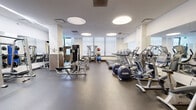 Located at 317 North Broad Street in Philadelphia, this fitness center offers a well-appointed exercise space designed to accommodate a wide range of workout needs. The room features a clean, modern aesthetic with white and warm yellow walls, rubber flooring, and a bright atmosphere enhanced by distinctive circular recessed ceiling lights and large windows that allow natural light to fill the space.

The cardio section is equipped with multiple treadmills, elliptical trainers, and stationary bikes, all arranged to provide ample room for movement. Strength training is well-represented with a cable and pulley machine system, a squat/power rack, a flat weight bench, and a full dumbbell rack spanning a wide range of weights. A barbell and weight plates area, along with medicine balls and a plyo box, cater to functional and dynamic training.

Floor-to-ceiling mirrors line multiple walls, amplifying the sense of space and allowing users to monitor their form. A wall-mounted flat-screen television adds an entertainment element. The entrance features elegant wood-framed French doors that provide a warm contrast to the otherwise contemporary interior. A water fountain is conveniently positioned near the entrance. The overall layout is open and spacious, offering a professional and functional environment well-suited for a comprehensive fitness routine.