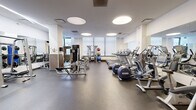 Located at 317 North Broad Street in Philadelphia, this fitness center offers a well-appointed exercise space designed to accommodate a wide range of workout needs. The room features a clean, modern aesthetic with white and warm yellow walls, rubber flooring, and a bright atmosphere enhanced by distinctive circular recessed ceiling lights and large windows that allow natural light to fill the space.

The cardio section is equipped with multiple treadmills, elliptical trainers, and stationary bikes, all arranged to provide ample room for movement. Strength training is well-represented with a cable and pulley machine system, a squat/power rack, a flat weight bench, and a full dumbbell rack spanning a wide range of weights. A barbell and weight plates area, along with medicine balls and a plyo box, cater to functional and dynamic training.

Floor-to-ceiling mirrors line multiple walls, amplifying the sense of space and allowing users to monitor their form. A wall-mounted flat-screen television adds an entertainment element. The entrance features elegant wood-framed French doors that provide a warm contrast to the otherwise contemporary interior. A water fountain is conveniently positioned near the entrance. The overall layout is open and spacious, offering a professional and functional environment well-suited for a comprehensive fitness routine.