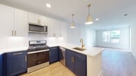 This single-floor residence at Alta Porter On Peachtree in Brookhaven, Georgia, offers a thoughtfully designed open-concept layout with modern finishes throughout. The kitchen serves as a central highlight, featuring a striking two-tone cabinet design with navy lower cabinets and crisp white upper cabinets, all accented by gold hardware. White quartz countertops extend across the peninsula, which includes an undermount sink with a gold faucet and is illuminated by stylish pendant lighting. Stainless steel appliances, including a gas range, microwave, dishwasher, and refrigerator, complete the well-equipped space. A white subway tile backsplash adds a clean, polished touch.

The living area flows seamlessly from the kitchen, featuring light hardwood-style flooring and a ceiling fan that enhances the bright, airy atmosphere. A large window allows natural light to fill the space, creating an inviting ambiance. In-unit stacked laundry is conveniently accessible near the main living area.

The bathroom features a glass-enclosed walk-in shower with large white tiles and matte black hardware, complemented by light wood-style flooring. A generously sized walk-in closet with wire shelving and carpeted flooring provides ample storage. The overall design palette of soft whites, natural wood tones, and gold and black accents gives the space a cohesive, contemporary aesthetic.