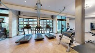 This well-appointed fitness center in Lakewood, Colorado offers a comprehensive workout environment across a generously sized single-floor layout. The main exercise area is thoughtfully designed with a herringbone-patterned hardwood floor that lends a refined, contemporary aesthetic throughout the space. Floor-to-ceiling glass garage-style doors and expansive windows flood the interior with natural light and provide open views of the surrounding greenery, creating an energizing atmosphere.

The cardio zone features a row of modern treadmills and elliptical machines positioned directly in front of the large windows, maximizing the connection to the outdoors. Adjacent to this area, a dedicated strength training section includes a multi-station cable machine, free weights, weight benches, and assorted resistance equipment, all set against full-length mirrors that visually expand the space.

A separate cycling studio accommodates multiple stationary bikes and is adorned with large framed mirrors, elegant sputnik-style pendant lighting, and clean white textured walls. An additional flex room serves as a yoga and functional training area, featuring stability balls, a BOSU trainer, a wall-mounted display screen, and a striking large-scale artistic mural that adds a dramatic, motivating focal point. The overall design balances functionality with a polished, upscale ambiance.