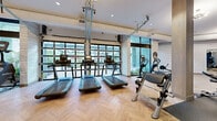 This well-appointed fitness center in Lakewood, Colorado offers a comprehensive workout environment across a generously sized single-floor layout. The main exercise area is thoughtfully designed with a herringbone-patterned hardwood floor that lends a refined, contemporary aesthetic throughout the space. Floor-to-ceiling glass garage-style doors and expansive windows flood the interior with natural light and provide open views of the surrounding greenery, creating an energizing atmosphere.

The cardio zone features a row of modern treadmills and elliptical machines positioned directly in front of the large windows, maximizing the connection to the outdoors. Adjacent to this area, a dedicated strength training section includes a multi-station cable machine, free weights, weight benches, and assorted resistance equipment, all set against full-length mirrors that visually expand the space.

A separate cycling studio accommodates multiple stationary bikes and is adorned with large framed mirrors, elegant sputnik-style pendant lighting, and clean white textured walls. An additional flex room serves as a yoga and functional training area, featuring stability balls, a BOSU trainer, a wall-mounted display screen, and a striking large-scale artistic mural that adds a dramatic, motivating focal point. The overall design balances functionality with a polished, upscale ambiance.