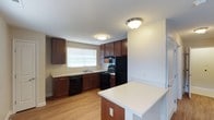 This single-floor residence offers a thoughtfully designed layout centered around a functional and attractive kitchen. The kitchen is outfitted with rich dark wood cabinetry that provides generous storage, complemented by light-toned countertops that create a pleasing contrast. Black appliances, including a refrigerator, electric range, and dishwasher, lend a sleek, cohesive look to the space. A peninsula-style breakfast bar extends from the cabinetry, offering a convenient spot for casual dining or meal prep. Warm hardwood-style flooring runs throughout, adding a sense of continuity and warmth to the interior. A window above the sink invites natural light into the kitchen, brightening the workspace and enhancing the overall ambiance. Flush-mount ceiling light fixtures provide consistent overhead illumination. The neutral wall tones work harmoniously with the cabinetry and flooring, creating a clean, inviting atmosphere. The open sightlines within the space contribute to an airy, accessible feel that makes everyday living both comfortable and practical.
