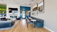 Located in Phoenix, AZ, this thoughtfully designed single-floor community clubroom offers a well-appointed gathering space that seamlessly blends social and relaxation areas. The open layout is anchored by light-toned hardwood flooring throughout, creating a warm and cohesive aesthetic.

The kitchen area is a standout feature, showcasing sleek white cabinetry with brushed hardware, white quartz countertops, a large central island with bar seating, and a striking black-framed grid window that floods the space with natural light. A living green wall with a playful neon "Drinks on Us" sign adds a vibrant focal point, complemented by elegant glass pendant lighting overhead.

The living area centers around a stone-accented fireplace with a large wall-mounted TV, flanked by a lounge chair and a plush sectional sofa set atop a textured area rug. A foosball table adds a recreational element nearby. The lounge dining zone features a deep teal channeled banquette, small round tables, and modern side chairs beneath pendant lights, with Arizona-themed artwork adorning the walls.

Bold teal accent walls contrast beautifully against neutral tones throughout, adding visual depth. Expansive sliding glass doors open directly to an outdoor pool terrace, bathing the interior in natural light and offering seamless indoor-outdoor connectivity.