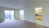 This single-floor residence features a thoughtfully arranged layout that maximizes openness and functionality. The main living area is generously proportioned, finished with wall-to-wall carpeting in neutral tones that lend a clean, understated aesthetic. White walls and standard-height ceilings keep the space feeling bright and airy.

A sliding glass door along one wall opens directly to a balcony enclosed by a metal railing, allowing natural light to flow into the living space while providing an outdoor retreat. An adjacent room, visible through an open passageway, features a window that adds further natural illumination.

To the right, the kitchen is accessible via an open entryway and is equipped with light wood cabinetry, a gas range, and a dishwasher. The kitchen features tile or hard flooring, contrasting with the carpeted living areas. Under-cabinet lighting and an efficient layout make the kitchen both practical and well-organized.

Overall, the space offers a functional, straightforward design with consistent neutral tones, practical finishes, and good access to natural light, making it a versatile and livable single-floor residence.