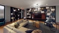 This elegantly designed single-floor space offers a refined and cohesive lounge environment that blends contemporary aesthetics with warm, natural materials. The distinctive cork flooring runs throughout, providing both visual warmth and a unique textural quality underfoot. Floor-to-ceiling windows with sheer roller shades line multiple walls, allowing abundant natural light to permeate the interior while maintaining a soft, diffused ambiance.

The space is organized into distinct seating zones, each thoughtfully curated. One area features a generous sectional sofa in neutral tones, accented with decorative pillows and anchored by a dark area rug. Natural wood stump side tables add an organic touch. A second lounge zone showcases tan leather sofas arranged around a glass coffee table, bathed in light from the expansive corner windows.

A dramatic built-in shelving wall, finished in dark tones with integrated lighting, serves as both a functional and decorative focal point. It frames a large flat-screen television and displays curated décor and books. A contemporary globe chandelier adds a striking overhead accent.

A long corridor lined with additional illuminated built-in shelving and glass-fronted rooms connects the various zones, reinforcing the space's open, flowing layout. The overall atmosphere is sophisticated, calm, and well-appointed.