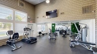 Located at 1820 Hickory Creek Ln in Columbus, Ohio, this fitness center offers a thoughtfully designed workout space that accommodates a comprehensive range of exercise options. The facility is defined by its soaring ceilings and shingle-style exterior walls that rise dramatically upward, lending the interior a sense of openness and volume. Large windows line the walls, allowing abundant natural light to fill the space and providing views of the surrounding outdoor greenery. The durable rubber-speckle flooring throughout provides both safety and comfort for all types of workouts. Cardio equipment is well represented, with multiple treadmills, an elliptical, a stationary bike, a rowing machine, and a stair climber available for use. Strength training needs are met with a cable/pulley multi-station machine, adjustable benches, a dedicated free weights area with a full dumbbell rack and kettlebells, and various selectorized machines. A vivid green moss wall accent adds a natural, modern aesthetic touch, complemented by large wall-mounted mirrors that enhance the sense of space. Mounted flat-screen televisions and a ceiling fan contribute to a comfortable and engaging workout environment. A water fountain station is also conveniently available. Recessed lighting and an exit door with direct outdoor access round out this functional and well-maintained community fitness center.