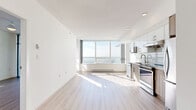 This thoughtfully designed one-bedroom, one-bathroom unit offers a clean, modern aesthetic defined by an all-white palette, light-toned wide-plank laminate flooring, and an abundance of natural light. The open-concept living and kitchen area is anchored by a dramatic wall of floor-to-ceiling windows, framing expansive panoramic views that flood the space with daylight and create a sense of openness well beyond the unit's compact footprint.

The kitchen is efficiently designed with a two-tone cabinetry scheme — crisp white upper cabinets paired with warm wood-grain lower cabinets — complemented by a white subway tile backsplash and light quartz countertops. A full suite of stainless steel appliances, including a built-in microwave, electric range, dishwasher, and bottom-mount refrigerator, provides complete functionality.

The bedroom is a well-proportioned, serene retreat with access to a private balcony through a full-height sliding glass door, allowing natural light to pour in. Baseboard heating ensures year-round comfort.

The bathroom is finished in a clean, spa-like style featuring large-format white wall tiles, a soaker tub with overhead shower, a sleek vanity with stone countertop, and a large mirror. Recessed lighting and textured flooring complete the polished look. Overall, the unit presents a bright, contemporary living environment with a cohesive modern design language.