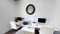 This single-floor space functions as a well-appointed business center designed to support productivity and convenience. The room features a clean, modern aesthetic with white walls that create a bright and open atmosphere. At the center of the space sits a streamlined white workstation that accommodates two computer setups, each equipped with desktop monitors and adjustable task lighting to support extended work sessions. A multifunction inkjet printer and a laser printer are positioned on the desk surface, ensuring a full range of document handling capabilities. White ergonomic chairs provide comfortable seating at each workstation. The flooring features a checkered tile pattern near the workstation area, transitioning to carpeted flooring, adding visual interest to the layout. A bold circular mirror with a dark ridged frame serves as a focal decorative element on the wall above the desk, lending the space a touch of contemporary style. A small potted plant with vibrant red blooms adds a natural accent and warmth to the otherwise crisp, monochromatic color scheme. The overall ambiance is professional and functional, with a tidy arrangement that makes efficient use of the available space, making it well-suited for tasks such as printing, computing, and general business needs.