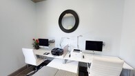 This single-floor space functions as a well-appointed business center designed to support productivity and convenience. The room features a clean, modern aesthetic with white walls that create a bright and open atmosphere. At the center of the space sits a streamlined white workstation that accommodates two computer setups, each equipped with desktop monitors and adjustable task lighting to support extended work sessions. A multifunction inkjet printer and a laser printer are positioned on the desk surface, ensuring a full range of document handling capabilities. White ergonomic chairs provide comfortable seating at each workstation. The flooring features a checkered tile pattern near the workstation area, transitioning to carpeted flooring, adding visual interest to the layout. A bold circular mirror with a dark ridged frame serves as a focal decorative element on the wall above the desk, lending the space a touch of contemporary style. A small potted plant with vibrant red blooms adds a natural accent and warmth to the otherwise crisp, monochromatic color scheme. The overall ambiance is professional and functional, with a tidy arrangement that makes efficient use of the available space, making it well-suited for tasks such as printing, computing, and general business needs.