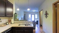 This single-floor space showcases an open-concept layout where the kitchen and living area connect seamlessly, creating a cohesive and functional environment. The kitchen features dark espresso cabinetry paired with light countertops, a double-basin stainless steel sink, and granite-style backsplash detailing that adds a touch of elegance. Two pendant lights hang above the kitchen island or counter overhang, providing both task lighting and a decorative focal point that draws the eye upward toward the smooth, well-lit ceiling.

The open sightline from the kitchen extends directly into the living area, where a gray sofa anchors the space. Natural light enters through a set of front-facing windows flanking what appears to be an entry door, brightening the interior and creating an inviting atmosphere. Warm hardwood-style flooring runs continuously throughout, unifying the kitchen and living spaces. A hallway visible to the right suggests additional rooms beyond, with a doorway offering access to further private areas of the home. Overall, the space presents a clean, modern aesthetic with practical design choices suited for everyday living.