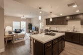 Located at 26000 W Lugonia Ave in Redlands, California, this single-story residence offers a well-designed open-concept layout with contemporary finishes throughout. The kitchen is a standout feature, showcasing rich dark espresso cabinetry, quartz countertops, a large center island with pendant lighting, and stainless steel appliances including a refrigerator, gas range, microwave, and dishwasher. Wood-look flooring flows seamlessly through the kitchen and into the living area, which is bathed in natural light from a sliding glass door that opens to an outdoor balcony. The living space provides a generous, comfortable area ideal for both relaxing and entertaining. Two well-appointed bedrooms feature plush carpeting, natural light from large windows with blinds, and neutral warm-toned walls that create a calm, restful ambiance. The two bathrooms are finished with dark cabinetry, quartz countertops, and wood-look flooring; one features a tub-shower combination, while the other offers a sleek, streamlined design. A built-in desk area in the kitchen adds functional versatility. Throughout the residence, recessed lighting and pendant fixtures contribute to a bright, modern atmosphere that is both stylish and practical.