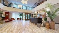 Located at 111 E Broad St in Falls Church, Virginia, this expansive lobby hallway makes a striking first impression with its soaring double-height atrium ceiling and an open mezzanine level framed by a sleek black metal railing above. The floor is laid in a classic herringbone pattern of warm-toned marble tile, lending the space a refined, timeless character. A cluster of large globe pendant lights cascades from the ceiling, providing soft, ambient illumination throughout. At the center of the lobby, a dark-finished reception desk serves as the focal point, flanked by lush tropical plants that bring life and warmth to the environment. Seating areas are thoughtfully arranged throughout, featuring rich terracotta-orange velvet sofas, tan leather lounge chairs, and small round accent tables, all evoking a sophisticated mid-century modern aesthetic. Floor-to-ceiling glass entry doors along one wall allow abundant natural light to filter in, while sheer white curtains diffuse the light softly across the interior. Framed artwork and decorative wall sconces accent the elevator alcove, adding a curated, boutique feel. The overall ambiance is welcoming and upscale, balancing open, airy architecture with warm, carefully selected furnishings.