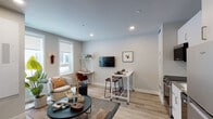 This single-floor unit is designed with an efficient, modern layout that makes excellent use of every square foot. The open-concept living area welcomes natural light through multiple windows fitted with roller shades, illuminating light gray walls and warm light wood flooring that run throughout the space. Recessed lighting complements the natural light, creating a bright and inviting ambiance. The living area offers a comfortable lounge space alongside a compact dining setup with a mobile bar-height table and stools, with a wall-mounted TV completing the functional arrangement.

The kitchen is outfitted with full stainless steel appliances, including a range, microwave, dishwasher, and refrigerator. Flat-panel white cabinetry, a white penny tile backsplash, and a stainless steel sink contribute to a clean, contemporary aesthetic.

The bedroom is thoughtfully designed with a built-in storage system featuring overhead cabinets and open shelving that surround the bed, maximizing storage without sacrificing style.

The bathroom is finished with white subway tile in the walk-in shower, a floating vanity, and a large backlit mirror that enhances the sense of space. Every detail in this unit reflects a modern, well-considered design approach.