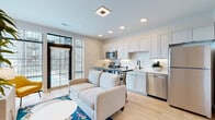 Located at 300 E Fifth Ave in Columbus, Ohio, this single-floor residence offers a thoughtfully designed open-concept layout with contemporary finishes throughout. Light hardwood flooring flows seamlessly from the entry hallway into the main living area, creating a bright and cohesive aesthetic.

The kitchen is a standout feature, equipped with stainless steel appliances including a range, microwave, dishwasher, and refrigerator. Shaker-style cabinetry in a soft gray tone lines the walls from floor to ceiling, complemented by white subway tile backsplash and clean white countertops. A built-in bar-height table with seating serves as both a dining and prep space, connecting the kitchen directly to the living area.

The living space is well-lit with recessed lighting and benefits from large windows that invite ample natural light. A spacious hallway with recessed lighting leads from the entryway toward the living area, offering a welcoming sense of arrival.

The bedroom is comfortably sized and includes a closet with a stacked in-unit washer and dryer for added convenience. The bathroom features a walk-in glass-enclosed shower with white tile surround and matte black fixtures, a single-sink vanity with stone countertop, and light wood-tone flooring that ties the space together with a clean, modern feel.