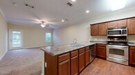 This two-story home is thoughtfully laid out across two levels connected by a carpeted staircase with dual handrails. The main level centers around an open-concept kitchen and living area. The kitchen is well-appointed with rich medium-toned wood cabinetry, granite-style countertops, stainless steel appliances including a range, built-in microwave, and refrigerator, plus a dishwasher. A central island with an integrated double sink and additional cabinetry extends toward the adjoining living area, which features hardwood-style flooring that transitions to carpet. Crown molding, recessed lighting, and a ceiling fan add polish to the shared space, which benefits from natural light through a window. An attached garage with an automatic opener and interior access door provides convenient entry. Upstairs, the primary suite is generously proportioned, featuring plush carpet, a ceiling fan, and multiple windows. The en-suite bath offers a dual-sink vanity with ample cabinetry, tile flooring, and direct access to a walk-in closet with built-in shelving. Additional carpeted rooms provide flexible living space throughout the upper level.
