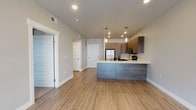 This contemporary single-floor unit showcases a thoughtfully designed open-concept layout that seamlessly connects the kitchen and living areas. The kitchen is a standout feature, outfitted with dark wood-grain cabinetry extending from floor to ceiling, sleek white quartz countertops, and a full suite of stainless steel appliances including a refrigerator, electric range, and over-the-range microwave. A peninsula with an undermount sink and stylish pendant lighting serves as both a functional workspace and a natural divider between the kitchen and the expansive living area. Light wood-look flooring flows throughout the main living spaces, complementing the neutral, light gray walls and recessed lighting that give the interior a bright, airy feel. The spacious living area benefits from a large floor-to-ceiling window with roller shades, allowing generous natural light and outdoor views. The bedroom features plush carpeting and a window with horizontal blinds, providing a quiet and comfortable retreat. The bathroom is finished with wood-toned cabinetry, white quartz countertops, a large framed mirror with vanity lighting, and a tub-shower combination, all unified by consistent wood-look flooring that ties the space together cohesively.