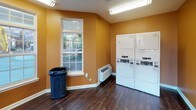 This single-floor shared laundry facility is designed with both functionality and comfort in mind. The space is characterized by warm amber-toned walls and dark hardwood-style flooring that create a welcoming atmosphere uncommon in utility spaces. Natural light flows generously throughout the room via multiple double-hung windows with white trim, offering views of surrounding greenery and neighboring structures. A glass-paneled exterior door provides additional brightness and direct outdoor access.

The laundry area is well-equipped, featuring a row of top-loading washing machines along one wall and stacked dryer units positioned nearby, offering ample capacity for shared use. A payment terminal is conveniently mounted near the washers. The facility also includes a large folding table supported by sleek hairpin legs, providing a practical surface for sorting and folding laundry. A wall-mounted fire extinguisher and a waste bin are thoughtfully placed throughout for safety and cleanliness.

Overhead fluorescent lighting supplements the natural light, ensuring the space remains bright at all hours. A wall-mounted HVAC unit maintains comfortable temperatures year-round. The overall layout is open and efficient, making this a practical and well-appointed shared laundry facility.
