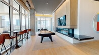 This single-floor amenity space is thoughtfully designed with a contemporary aesthetic, combining high-end recreational features with refined architectural details. The entry corridor introduces the space with large-format tile flooring, crisp white walls displaying curated framed artwork, and clean recessed lighting that sets a polished tone throughout.

The main lounge area is a standout, showcasing light hardwood flooring, a sleek billiards table centered on a neutral area rug, and a dramatic wall-mounted linear gas fireplace framed in dark stone beneath warm wood cabinetry and a large flat-screen television. Ambient cove lighting along the ceiling adds a warm, sophisticated glow. Floor-to-ceiling glass walls flood the space with natural light and offer sweeping urban skyline views, while a bar-height seating area provides a social gathering spot alongside the glazed facade.

Adjacent to the lounge, a dedicated golf simulator room features turf flooring and a full projection screen, offering an immersive experience. A separate lounge alcove presents a bold oversized art installation above a tan leather sofa, contributing to the space's curated gallery-like character. Throughout, the design seamlessly blends entertainment, comfort, and contemporary style.