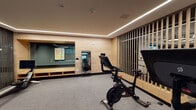 This contemporary fitness center is thoughtfully designed to accommodate a wide range of workout styles and fitness goals. The space is organized into several distinct zones, each with its own character and purpose. The main cardio area features rows of treadmills, ellipticals, and stair climbers positioned alongside expansive floor-to-ceiling windows that flood the space with natural light and offer views of the surrounding urban environment. A dedicated strength training zone provides a full complement of equipment, including cable machines, power racks with barbells, weight plates, and battle ropes, all set beneath an open, exposed ceiling with industrial-style black ductwork and modern accent lighting. A separate cycling studio houses multiple stationary bikes with individual screens, finished with warm wood-panel walls, recessed linear LED lighting, and a large mirror. Additional functional training space accommodates medicine balls, rowing machines, and open floor areas suitable for stretching or group exercise. Lush indoor plants are strategically placed throughout, adding a natural, calming element to the environment. Large sliding glass doors connect the interior to an outdoor terrace furnished with seating, extending the space and enhancing the overall sense of openness. The combination of modern materials, thoughtful lighting, and abundant natural light creates an energizing and inviting atmosphere.