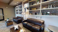 This single-floor space exudes a refined, library-inspired ambiance that seamlessly blends comfort with style. The focal point of the room is a striking floor-to-ceiling built-in bookshelf system, framed by exposed brick accent walls that lend an industrial yet warm character to the space. The shelves are thoughtfully arranged with books, decorative objects, and curated accents, adding depth and visual interest throughout.

The seating area is anchored by a pair of rich, dark brown leather sofas that invite relaxed conversation or quiet reading. A sleek floor lamp with multiple light heads provides both functional and ambient illumination, complementing the recessed ceiling lights that cast a soft, even glow across the room.

A patterned area rug grounds the seating arrangement, while a low coffee table with sculptural geometric decorative pieces adds an artistic touch. Original artwork is displayed prominently within the bookshelf alcove, contributing to the space's curated, gallery-like feel. The overall design strikes a balance between a classic reading lounge and a contemporary gathering space, making it both functional and visually compelling.