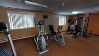 This fitness center is a well-equipped, single-floor exercise facility designed to meet a variety of workout needs. The space is divided into functional zones, each serving a distinct purpose for users. The main exercise area features a selection of cardio machines, including a Landice treadmill, an elliptical trainer, and a recumbent stationary bike, providing ample options for cardiovascular workouts. A multi-station cable weight machine rounds out the strength training offerings, accommodating a range of resistance exercises. The walls are painted in warm tan and earthy orange tones, complemented by patterned carpet flooring throughout. Fluorescent lighting illuminates the space evenly, while several windows fitted with vertical blinds allow natural light to filter in. Decorative potted plants are placed throughout, adding a touch of greenery to the environment. A wall-mounted air conditioning unit provides climate control. Adjacent to the main workout area, a separate section houses a snack vending machine and a Dr. Pepper beverage vending machine, offering convenient refreshment options. A small counter area with cabinetry and a coffee maker provides an additional amenity for visitors. The overall layout is practical and straightforward, making it easy to navigate between exercise and refreshment areas.