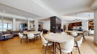 Located at 2274 Princess St in Kingston, Ontario, this single-floor amenity lounge offers a generously sized, open-concept layout designed for communal gathering and relaxation. The space is anchored by warm, wide-plank hardwood flooring laid in a herringbone pattern, crisp white ceilings with recessed lighting, and a sophisticated modern aesthetic throughout.

The kitchen area is a standout feature, equipped with rich walnut cabinetry, stainless steel appliances including a full-size refrigerator, a gas range, and a dishwasher. A striking dark-tiled island with a white countertop and bar seating serves as the kitchen's centerpiece, complemented by ambient open shelving and pendant lighting. A dining area adjacent to the kitchen features a round table and chairs bathed in natural light from large sliding glass doors that open to views of the surrounding grounds.

Multiple lounge zones provide versatile seating options: a U-shaped sectional sofa in rich leather anchors one relaxed lounge area, while a curved caramel leather sofa with marble accent tables defines a more intimate sitting space. Several round dining tables with white chairs are thoughtfully arranged throughout the open floor plan. A bold dark-tiled architectural column adds a dramatic focal point. Glass partition walls allow light to flow freely between zones, enhancing the bright, airy ambiance of the entire space.