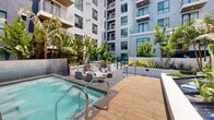 Located at 201 The Promenade N in downtown Long Beach, California, this outdoor amenity deck offers a thoughtfully designed resort-style retreat set within a modern multi-story residential building. The space centers around a sparkling spa pool with sleek steel entry rails, surrounded by warm wood-tone deck tiles that lend a polished, contemporary feel. Adjacent to the pool, a sunken lounge area features a curved sectional sofa, accent stools, and rattan chairs arranged for relaxed conversation and poolside enjoyment. Lush tropical landscaping defines the perimeter, with towering palm trees, banana leaf plants, agave, and a variety of tropical foliage creating a verdant, resort-inspired atmosphere. Raised planters with glass panel accents add dimension and visual interest throughout the deck. A standout feature is the recessed hammock nook, nestled among the greenery and cushioned with blue accent pillows, offering a quiet spot for relaxation. A water feature with a cascading wall element adds an ambient, tranquil quality to the environment. Overlooking the surrounding urban streetscape, the deck affords open-sky views and glimpses of neighboring downtown buildings, including a notable large-scale mural visible from the pool area. The overall ambiance balances urban energy with tropical serenity, making this a distinctive and inviting outdoor amenity.