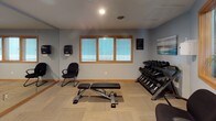 This single-story fitness and common area facility is thoughtfully designed to support an active lifestyle while maintaining a welcoming atmosphere throughout. The space is finished with cool blue-gray walls, warm wood trim accents, and light tile flooring in the common areas, creating a cohesive and calming aesthetic.

The entrance features a glass-paneled door flanked by sidelight windows with wood trim, allowing natural light to filter into the vestibule. A dark area rug anchors the entryway, and the open sightlines lead directly into the adjoining common corridor.

The hallway is finished with large-format tile flooring and features a dark wood credenza topped with informational brochures and reading materials, accented by a large nature-inspired triptych painting above. Warm overhead lighting illuminates the passage to additional rooms.

The fitness area is divided into two functional zones. One section houses Matrix-brand cardio equipment, including a treadmill and elliptical trainer, positioned beneath a textured ceiling with flush lighting. The adjacent strength training area includes a full dumbbell rack, an adjustable workout bench, a task chair, and a window providing natural light. Carpet tiles cover the floor, and coastal-themed artwork decorates the walls.