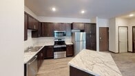 This single-floor residence offers a thoughtfully designed layout that balances functionality and comfort throughout its well-defined living spaces. The kitchen serves as a standout feature, showcasing rich dark-wood cabinetry, marble-patterned countertops, and a full suite of stainless steel appliances including a range, over-the-range microwave, dishwasher, and refrigerator. A central island with a matching marble-style surface provides additional workspace and casual dining potential. Light wood-look flooring flows through the kitchen and living area, lending warmth and continuity to the open layout. The living area benefits from a large sliding glass door dressed with vertical blinds, allowing abundant natural light and outdoor access. Two well-sized bedrooms feature soft wall-to-wall carpeting, neutral wall tones, and windows with horizontal blinds that frame views of surrounding greenery. The unit includes two full bathrooms, each equipped with a tub-shower combination, a dark-vanity with a white countertop, and classic mosaic or wood-look tile flooring. One bathroom is accessible directly through a walk-through closet area fitted with built-in shelving. Throughout the space, dark wood door and window trim provides a consistent design accent against the light, neutral walls, creating a clean and cohesive aesthetic.