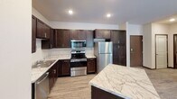 This single-floor residence offers a thoughtfully designed layout that balances functionality and comfort throughout its well-defined living spaces. The kitchen serves as a standout feature, showcasing rich dark-wood cabinetry, marble-patterned countertops, and a full suite of stainless steel appliances including a range, over-the-range microwave, dishwasher, and refrigerator. A central island with a matching marble-style surface provides additional workspace and casual dining potential. Light wood-look flooring flows through the kitchen and living area, lending warmth and continuity to the open layout. The living area benefits from a large sliding glass door dressed with vertical blinds, allowing abundant natural light and outdoor access. Two well-sized bedrooms feature soft wall-to-wall carpeting, neutral wall tones, and windows with horizontal blinds that frame views of surrounding greenery. The unit includes two full bathrooms, each equipped with a tub-shower combination, a dark-vanity with a white countertop, and classic mosaic or wood-look tile flooring. One bathroom is accessible directly through a walk-through closet area fitted with built-in shelving. Throughout the space, dark wood door and window trim provides a consistent design accent against the light, neutral walls, creating a clean and cohesive aesthetic.