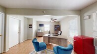 This single-floor office space is thoughtfully arranged to support day-to-day business operations in a welcoming and functional environment. The main area serves as both a reception and workstation zone, featuring a large wooden L-shaped desk equipped with a desktop computer, making it well-suited for client consultations and administrative tasks. A leather executive chair sits behind the desk, while two plush teal velvet accent chairs positioned on a decorative area rug offer comfortable seating for visitors. A wooden bookcase with posted notices and a wall-mounted flat-screen television add to the room's practical character. The space is finished with warm terracotta tile flooring throughout, contributing to a cohesive and easy-to-maintain interior. Natural light filters in through multiple windows dressed with horizontal blinds, and a ceiling fan promotes comfortable air circulation. A wall-mounted air conditioning unit provides additional climate control. A compact red retro-style mini refrigerator adds a distinctive visual accent. Adjacent to the main area, a secondary hallway leads to two interior doors, providing access to additional private rooms. Wall-mounted document organizers and a printer station round out the functional amenities, making this a well-equipped, professional space ready for active business use.