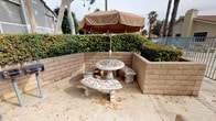 This outdoor communal space is thoughtfully designed to offer residents a variety of amenities for relaxation and recreation. At its heart is a dedicated picnic area nestled within a cinder block enclosure, featuring an ornate mosaic stone table and matching benches shaded by a large tan market umbrella — an inviting spot for casual outdoor dining. Adjacent to the seating area, a standalone barbecue grill and waste receptacle provide convenient options for outdoor cooking and entertaining.

A short path leads to the centerpiece of the outdoor amenities: a gated swimming pool enclosed by metal fencing for safety and privacy. The pool area is complemented by lounge seating and is framed by tall, stately palm trees that lend a warm, resort-like character to the setting.

Beyond the pool, the grounds open into a spacious landscaped common area featuring mature deciduous trees, manicured hedgerows, and winding concrete pathways that connect different areas of the property. Benches positioned along the walkways invite leisurely strolls or quiet moments outdoors. Multi-story residential buildings border the grounds, and the overall landscape conveys a well-established, community-oriented environment with a serene, park-like ambiance throughout.