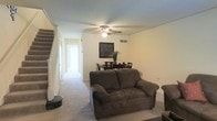 This two-story residence offers a functional and straightforward layout across its main and upper levels. The main floor living area is carpeted and opens into a connected dining space, creating a combined zone well-suited for everyday living and casual entertaining. A ceiling fan with integrated lighting is centrally mounted, providing both ventilation and ambient illumination throughout the shared space. Neutral-toned walls keep the interior feeling light and cohesive.

A staircase positioned along the left wall provides access to the upper floor, with a visible railing adding a structural accent to the room. A hallway extends toward the rear of the main floor, suggesting additional rooms or utility spaces beyond the immediate living and dining area. HVAC venting is visible along the upper wall, indicating climate control throughout the unit. The overall layout prioritizes practicality, with an open sightline from the living area toward the dining space, making the main floor feel connected and easy to navigate. The two-floor configuration adds separation between living and private areas of the home.
