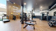 This fitness center is a thoughtfully designed workout facility that combines high-end aesthetics with comprehensive exercise equipment. The space is anchored by striking double-height ceilings, dramatic dark-painted structural columns, and a warm herringbone-patterned hardwood floor that runs throughout.

A standout design feature is the full-height wood slat accent wall adorned with illuminated motivational text — "Grind. Work. Burn. Focus." — serving as a bold focal point that sets the tone for the space. The walls feature a two-tone treatment with navy blue wainscoting below and crisp white above, lending a polished, contemporary feel.

Natural light pours in through expansive floor-to-ceiling windows and glass doors that open to an outdoor area, creating an airy and energizing atmosphere. A glass-railed mezzanine overlooks the main floor, adding architectural depth and visual interest.

The facility is outfitted with an extensive array of equipment, including multi-station cable and strength machines, TRX suspension trainers, adjustable benches, a full dumbbell rack, treadmills, stationary bikes, and cardio runners. A mirrored free weights area and a dedicated restroom further enhance the functionality of the space. Recessed lighting throughout ensures a well-lit environment at all hours, making this fitness center a complete and inviting destination for training.