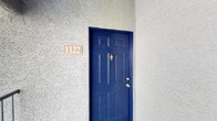 This single-floor unit is set within a well-maintained residential community characterized by lush green landscaping, mature trees, and winding pathways that create a welcoming outdoor environment. The exterior features stucco construction with a tile roof, chimneys, and balconies that contribute to its classic architectural character. A bold blue front door with brass hardware serves as a distinctive and inviting entryway.

Inside, the main living area is finished with plush wall-to-wall carpeting and benefits from vaulted ceilings that enhance the sense of openness and airiness. A ceiling fan with an integrated light fixture is centrally positioned overhead, providing both comfort and illumination. Natural light flows into the space through a large window and a glass-paneled door, which also offers direct access to the outdoors. The neutral wall tones and clean lines throughout create a bright, versatile interior ready to be personalized.

The surrounding community grounds are thoughtfully landscaped with grassy lawns, mature shade trees, and well-kept shrubs, offering a serene and visually appealing setting that complements the residential atmosphere of the property.