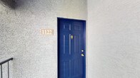 This single-floor unit is set within a well-maintained residential community characterized by lush green landscaping, mature trees, and winding pathways that create a welcoming outdoor environment. The exterior features stucco construction with a tile roof, chimneys, and balconies that contribute to its classic architectural character. A bold blue front door with brass hardware serves as a distinctive and inviting entryway.

Inside, the main living area is finished with plush wall-to-wall carpeting and benefits from vaulted ceilings that enhance the sense of openness and airiness. A ceiling fan with an integrated light fixture is centrally positioned overhead, providing both comfort and illumination. Natural light flows into the space through a large window and a glass-paneled door, which also offers direct access to the outdoors. The neutral wall tones and clean lines throughout create a bright, versatile interior ready to be personalized.

The surrounding community grounds are thoughtfully landscaped with grassy lawns, mature shade trees, and well-kept shrubs, offering a serene and visually appealing setting that complements the residential atmosphere of the property.