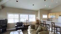 This single-floor space offers a warm and inviting atmosphere, thoughtfully designed to maximize comfort and functionality. The living area is a standout feature, boasting soaring vaulted ceilings fitted with recessed lighting that complement the abundant natural light streaming in through a generous row of windows dressed with white horizontal blinds. The neutral, warm-toned walls create a calm and cohesive backdrop throughout the space. The living room is generously proportioned, comfortably accommodating a plush, earth-toned sofa adorned with decorative pillows and a classic leather armchair, arranged around a sleek glass coffee table — ideal for relaxed gatherings. A charming Tiffany-style table lamp on a wooden side table adds a touch of character and warmth to the seating area. Adjacent to the living room, a compact dining nook features a wooden bar-height table and chairs, seamlessly integrated into the open layout. The space is finished with soft, neutral carpeting underfoot, enhancing the overall sense of comfort. With its airy ceilings, plentiful windows, and well-connected living and dining areas, this space strikes a welcoming balance between casual and functional living.
