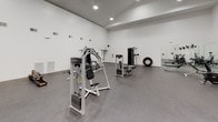 This fitness center offers a comprehensive and well-organized workout environment designed to accommodate a wide range of exercise routines. The space features soaring vaulted ceilings with recessed lighting, creating an open and airy atmosphere across the generously sized, single-floor facility. The floors are finished with durable rubber gym flooring, providing both safety and comfort during workouts.

The cardio area is equipped with multiple treadmills, upright and recumbent stationary bikes, and an elliptical trainer, all positioned in front of wall-mounted flat-screen televisions for an engaging workout experience. A large mirror is installed along one wall, adding depth to the space and supporting form-checking during exercise.

The strength training section features a variety of cable machines and multi-use weight stations from reputable brands, including Life Fitness equipment. Additional free-weight options are available, including a full rack of dumbbells. Functional training tools such as a tire and rowing machine round out the equipment offerings.

The facility is bright and clean, with crisp white walls, multiple ventilation units positioned high on the walls, and a sanitization station. Entry is through elegant French doors, and clearly marked exit signage ensures safety. The overall design is practical, spacious, and purpose-built for effective fitness training.