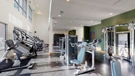 Located at 4600 Ross Ave in Dallas, TX, this modern fitness center is designed to provide a comprehensive workout experience within a spacious, single-floor layout. The facility boasts soaring ceilings and an abundance of natural light streaming through large floor-to-ceiling windows, offering pleasant views of the surrounding outdoor greenery. The polished concrete flooring runs throughout, lending a sleek, contemporary aesthetic that complements the clean white and bold green accent walls.

The cardio zone is well-appointed with a row of treadmills and elliptical machines, all positioned to take advantage of the natural light and views. A standout feature of the facility is the dedicated rock climbing wall, branded as the "Freedom Climber," which adds a dynamic and unique training option alongside a large flat-screen display for interactive workouts. The strength training area offers a comprehensive selection of cable machines, resistance equipment, and free weights, including a full dumbbell rack with wall-mounted mirrors.

A dedicated utility station features built-in blue cabinetry, a water fountain, and a hydration refill station, ensuring convenience during workouts. Ceiling fans provide additional airflow throughout the space. The overall ambiance is energetic yet functional, combining thoughtful design with a full suite of fitness amenities.