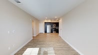 This single-floor space offers a clean, modern design centered around an open-concept layout that seamlessly connects the living area to the kitchen. The flooring throughout is a light hardwood-style material that adds warmth and continuity to the space, while recessed lighting in the ceiling provides bright, even illumination across the open floor plan.

The kitchen is a standout feature, outfitted with dark espresso cabinetry that creates a striking contrast against the neutral beige walls. Stainless steel appliances, including a refrigerator, range, and built-in microwave, lend a contemporary, professional feel. A tile backsplash adds a subtle decorative touch behind the cooking area. The kitchen layout is efficient and well-organized, with ample cabinetry for storage.

The living area is generously proportioned, offering a blank canvas with bright, neutral walls and clean white baseboards and trim. Natural light streams in, reflecting off the light-toned floors and enhancing the airy ambiance of the space. A doorway visible from the main living area suggests additional rooms beyond, contributing to a functional and versatile floor plan. Overall, the space conveys a fresh, move-in-ready aesthetic with thoughtful finishes throughout.