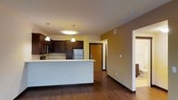 This single-floor space showcases a thoughtfully designed open-concept layout that seamlessly connects the kitchen and living areas. The kitchen is a standout feature, equipped with stainless steel appliances, including a full-size refrigerator, and a generous white countertop island that serves as both a workspace and a casual dining area. Dark espresso-toned cabinetry lines the walls, providing ample storage while adding a rich, warm contrast to the light-colored countertops and pale yellow walls. Pendant lighting hangs above the island, enhancing the functional ambiance with a touch of style. Rich hardwood flooring runs throughout the main living area, adding warmth and continuity to the space. Neutral warm tones on the walls contribute to an inviting and comfortable atmosphere. A natural wood-toned door near the kitchen leads to an adjacent area, suggesting additional rooms or a utility space beyond. The open hallway visible from the main area hints at further living quarters, maintaining a sense of spaciousness and flow throughout the floor plan. Overall, the space balances practicality with a cozy, welcoming aesthetic suited for everyday living.
